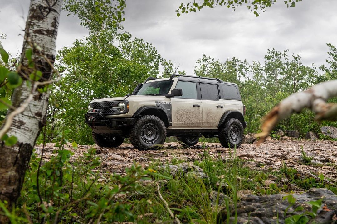 Tan Ford Bronco SUV on a rocky trail with green foliage and trees in the background, showcasing off-road capabilities and rugged design.