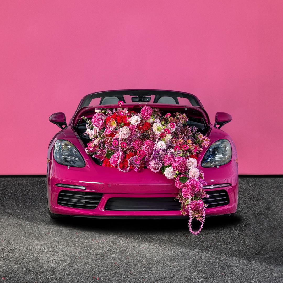 Hot pink convertible car filled with an array of colorful flowers on a grey surface in front of a matching pink wall.