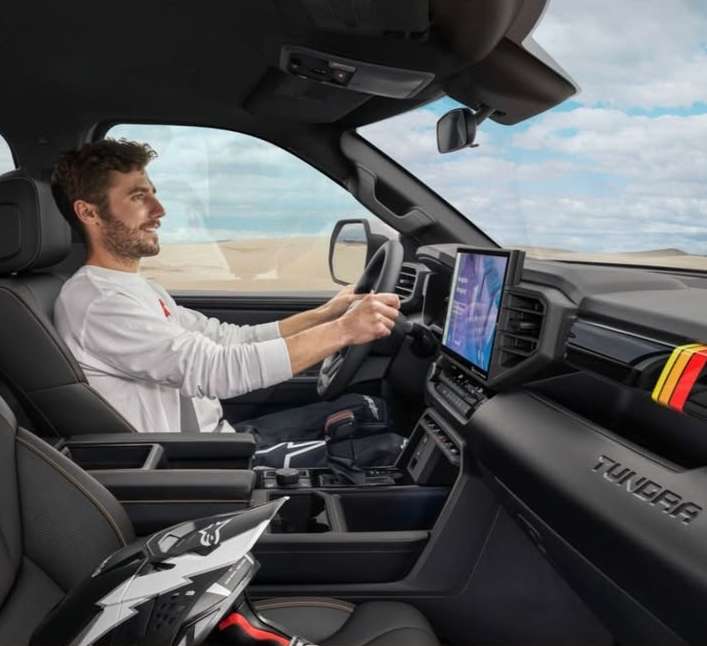 Man driving a modern pickup truck on a sunny day. The interior shows a helmet and digital displays against a desert landscape backdrop.
