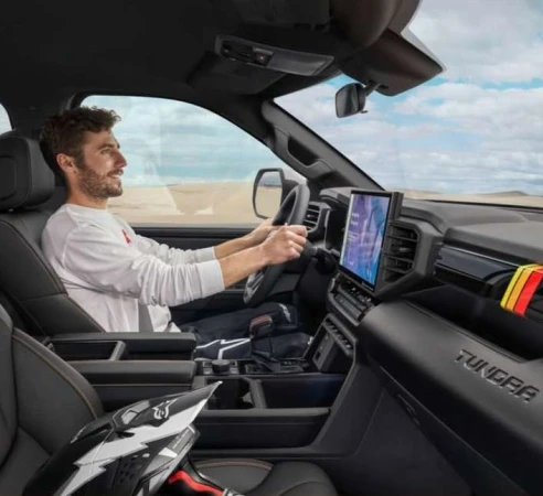 Man driving a modern pickup truck on a sunny day. The interior shows a helmet and digital displays against a desert landscape backdrop.
