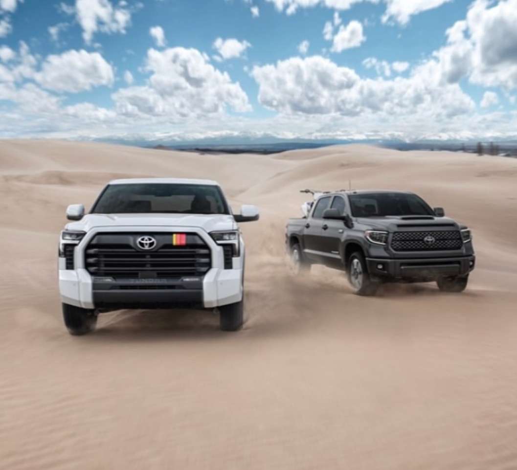Two Toyota Tundra pickup trucks, one white and one black, are driving on a sand dune under a blue sky with white clouds.