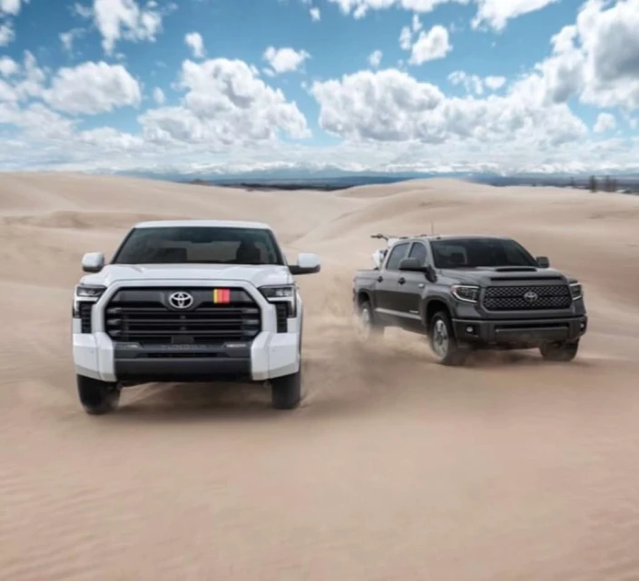 Two Toyota Tundra pickup trucks, one white and one black, are driving on a sand dune under a blue sky with white clouds.