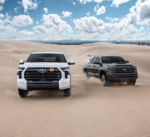 Two Toyota Tundra pickup trucks, one white and one black, are driving on a sand dune under a blue sky with white clouds.