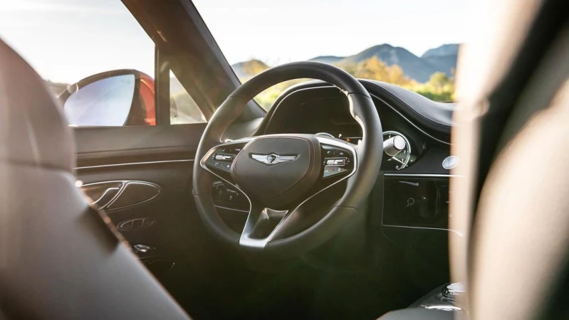 Interior view of a Genesis car with a black steering wheel, leather interior, and a dashboard against a scenic mountain backdrop.