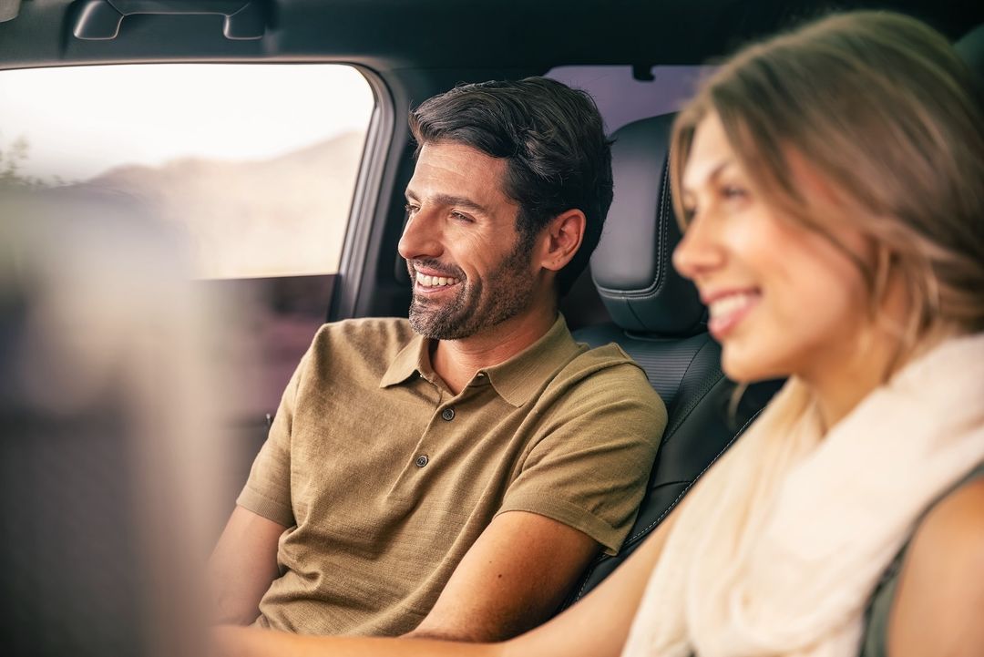 A smiling couple, an adult male and female, are seen inside a car with a blurred landscape visible through the window. They appear relaxed.