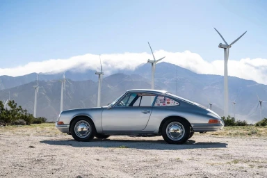 A vintage silver sports car parked on a gravel ground with numerous wind turbines and mountains under a clear sky in the background.