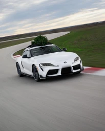 A white sports car with a small Christmas tree strapped to its roof speeds along an asphalt racetrack surrounded by grassy hills.