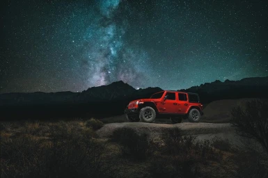 Red SUV parked on a dark rocky terrain beneath a vibrant starry night sky with the Milky Way visible.