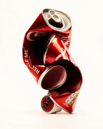 Crushed and distorted red Coca-Cola can on a white background, showing recycling information and a black bat logo on the bottom.