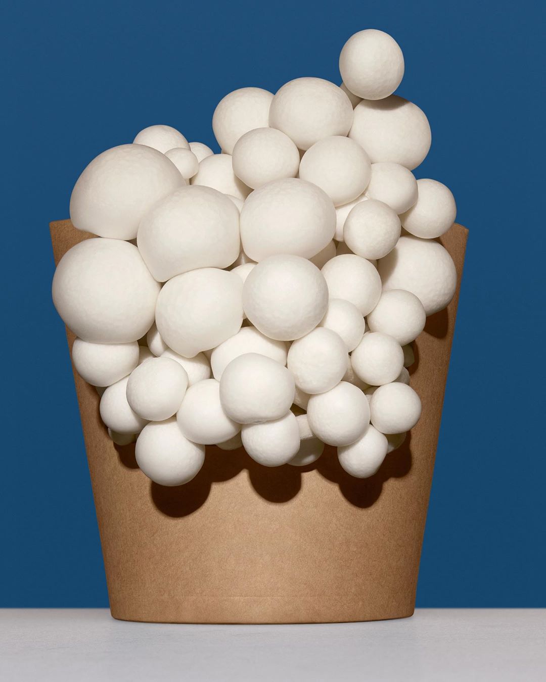Still life studio shot of white spherical mushrooms in a brown paper bag against a solid dark blue background creating a minimalist food still.