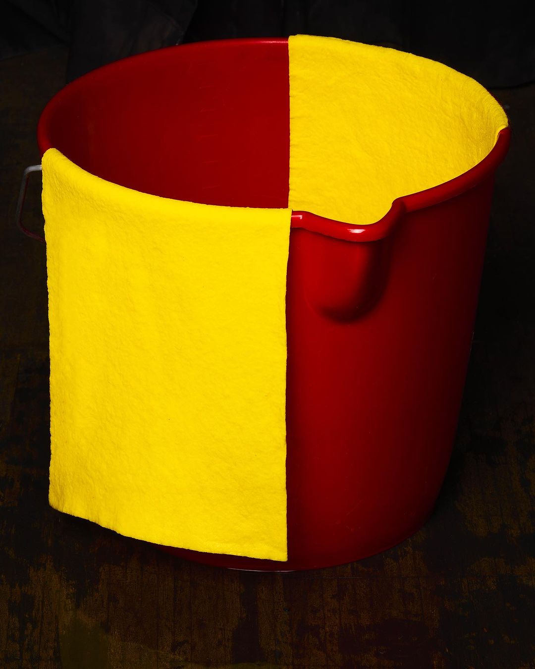 A red plastic bucket has a yellow cloth hanging from its side in a high-contrast studio shot, emphasizing color and texture.