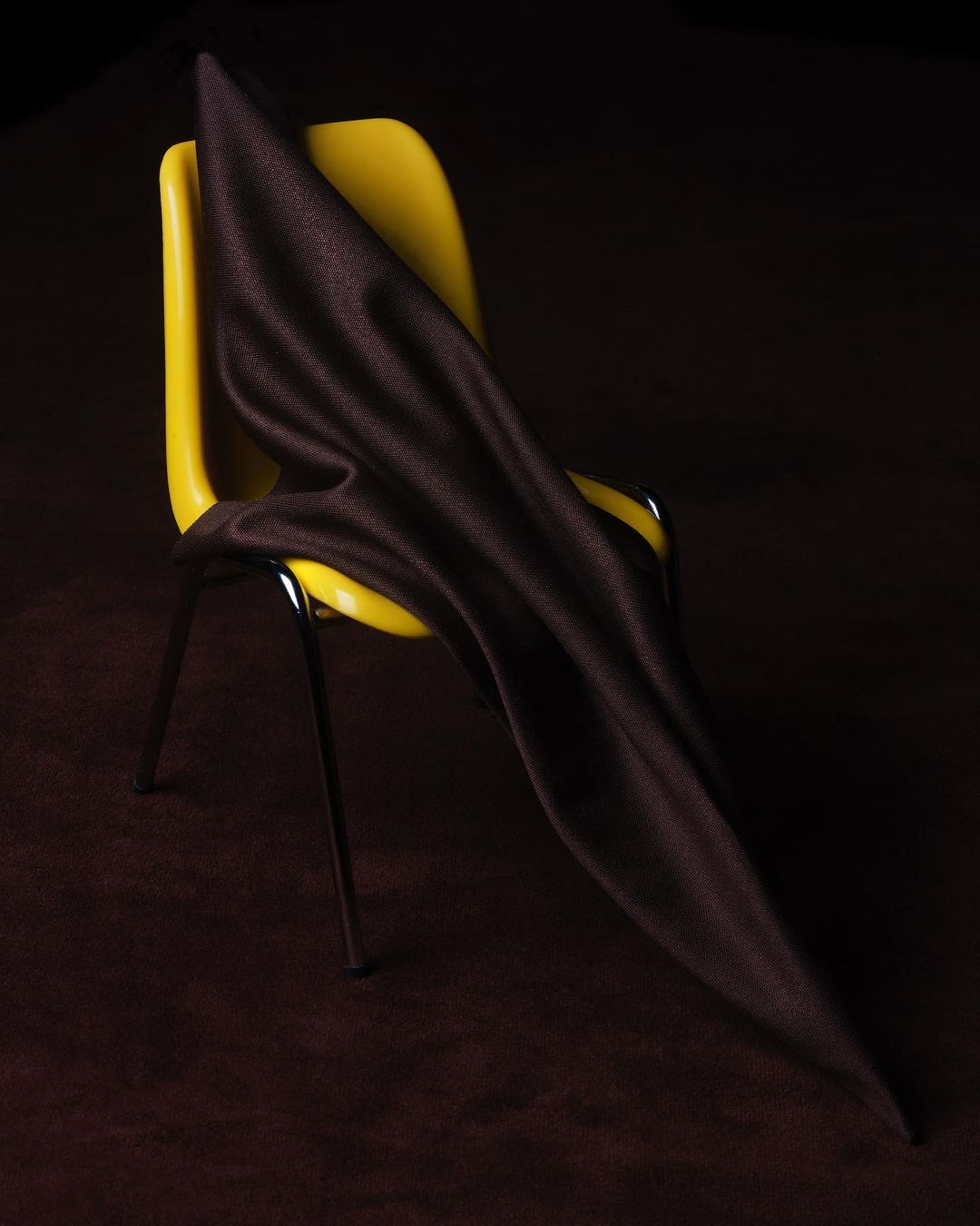 Dark brown textile draped on a yellow plastic chair, studio lighting, dark brown background, high angle and a slightly tilted view.