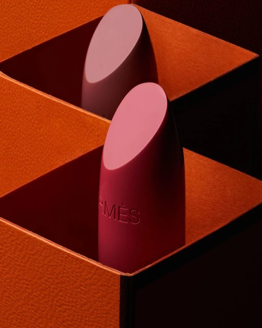 Two lipsticks are displayed in an orange box. The foreground lipstick is pink, and the background lipstick is light purple.