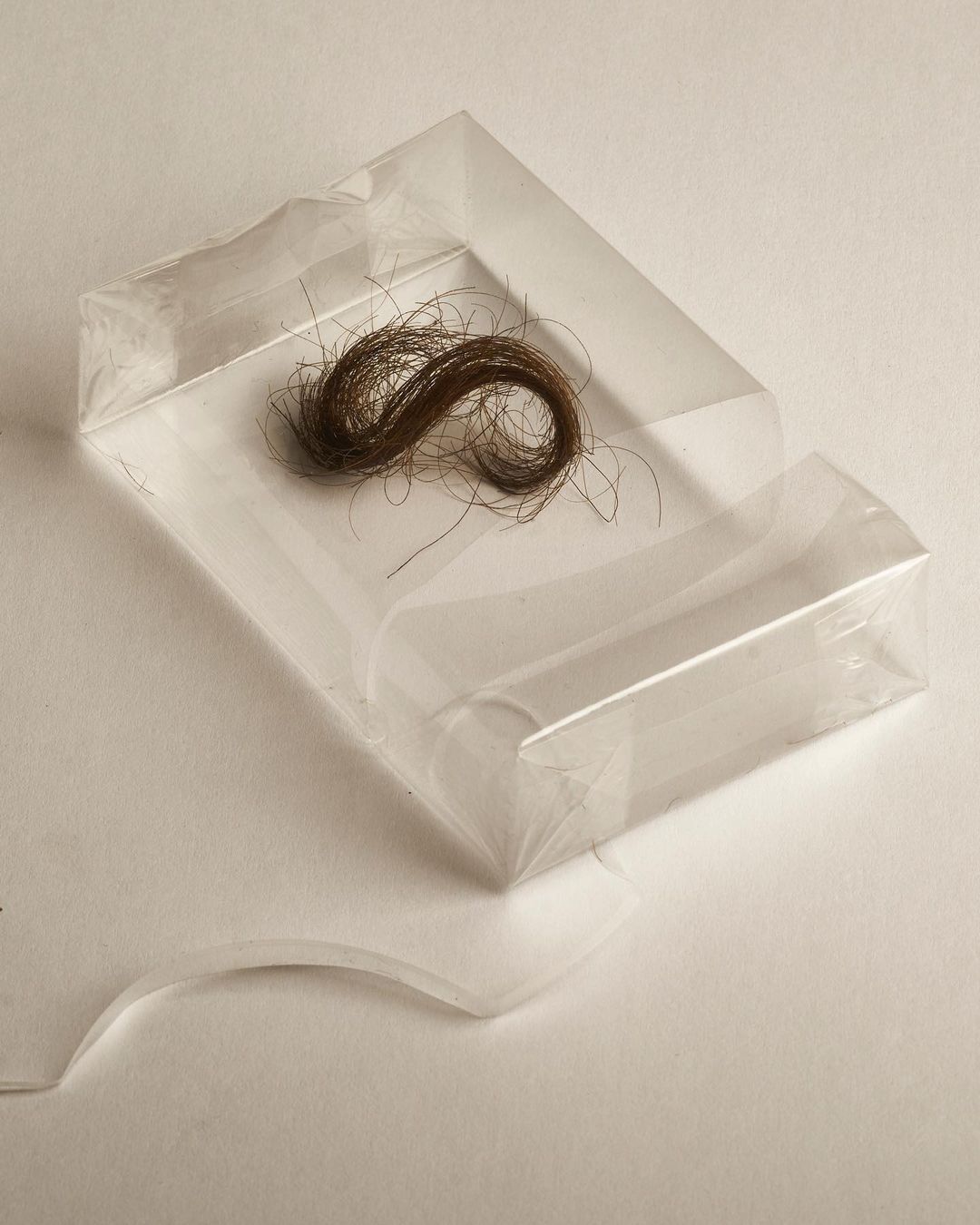 A lock of brown hair sits in a clear plastic case on a white surface, presented in a simple, close-up studio photograph.