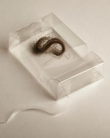 A lock of brown hair sits in a clear plastic case on a white surface, presented in a simple, close-up studio photograph.