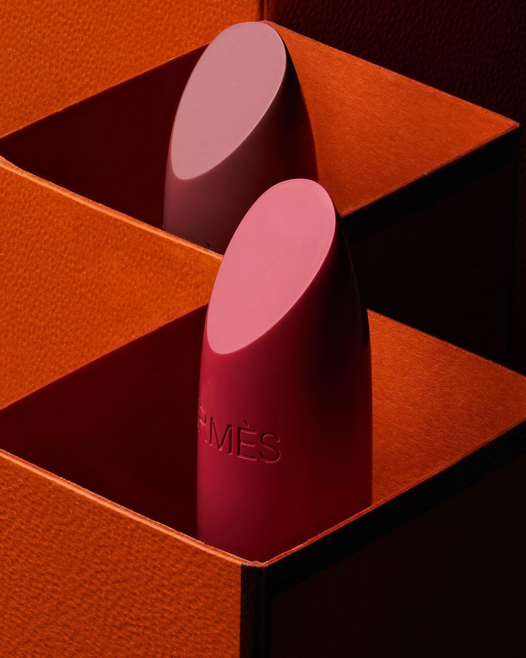 Two Hermès lipsticks are nestled inside orange packaging, the front lipstick displaying the Hermès logo, in a close-up studio shot.