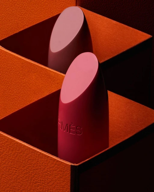 Two Hermès lipsticks are nestled inside orange packaging, the front lipstick displaying the Hermès logo, in a close-up studio shot.