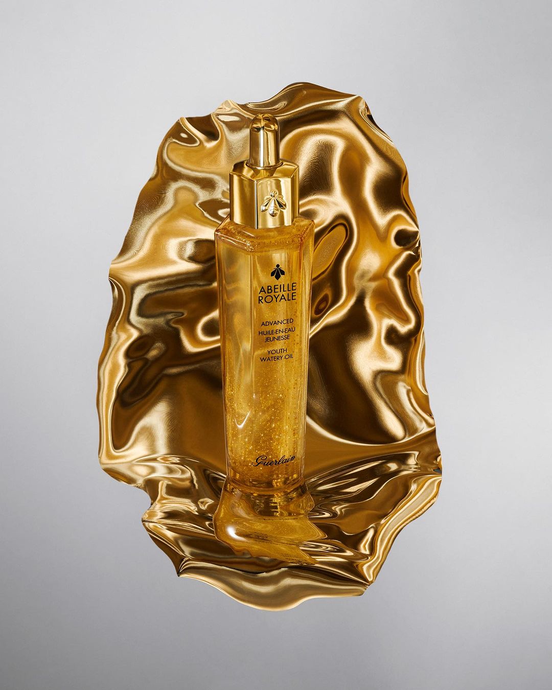Gold cosmetic serum bottle from Guerlain’s Abeille Royale line on a gold reflective surface against a gray backdrop, product shot.