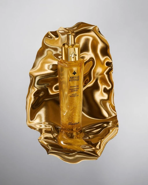 Gold cosmetic serum bottle from Guerlain’s Abeille Royale line on a gold reflective surface against a gray backdrop, product shot.