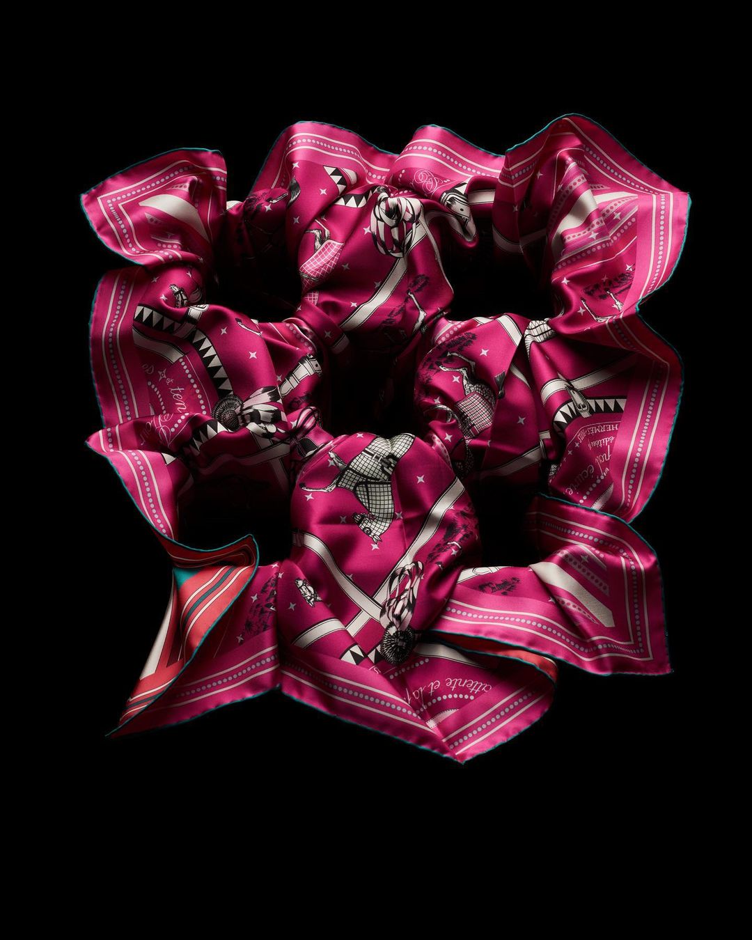 Close-up of a scrunchie made from a magenta and white patterned silk scarf, adorned with equestrian motifs, set against a stark black background.