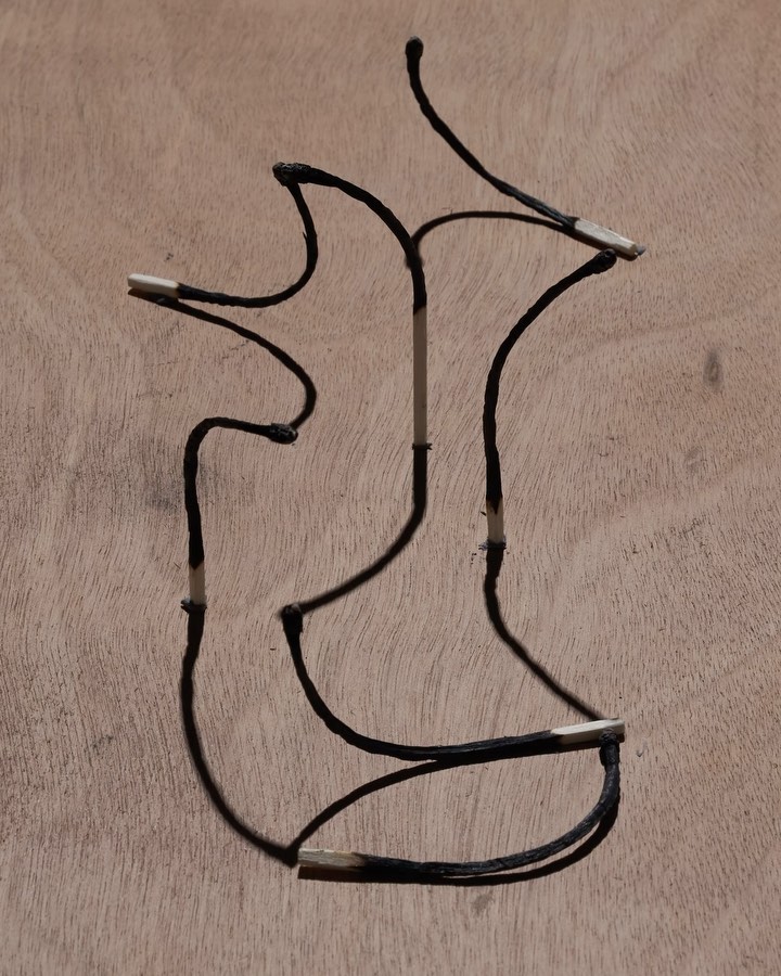 Burnt matchsticks arranged in an abstract shape against a light brown wooden surface, creating a visually striking contrast.