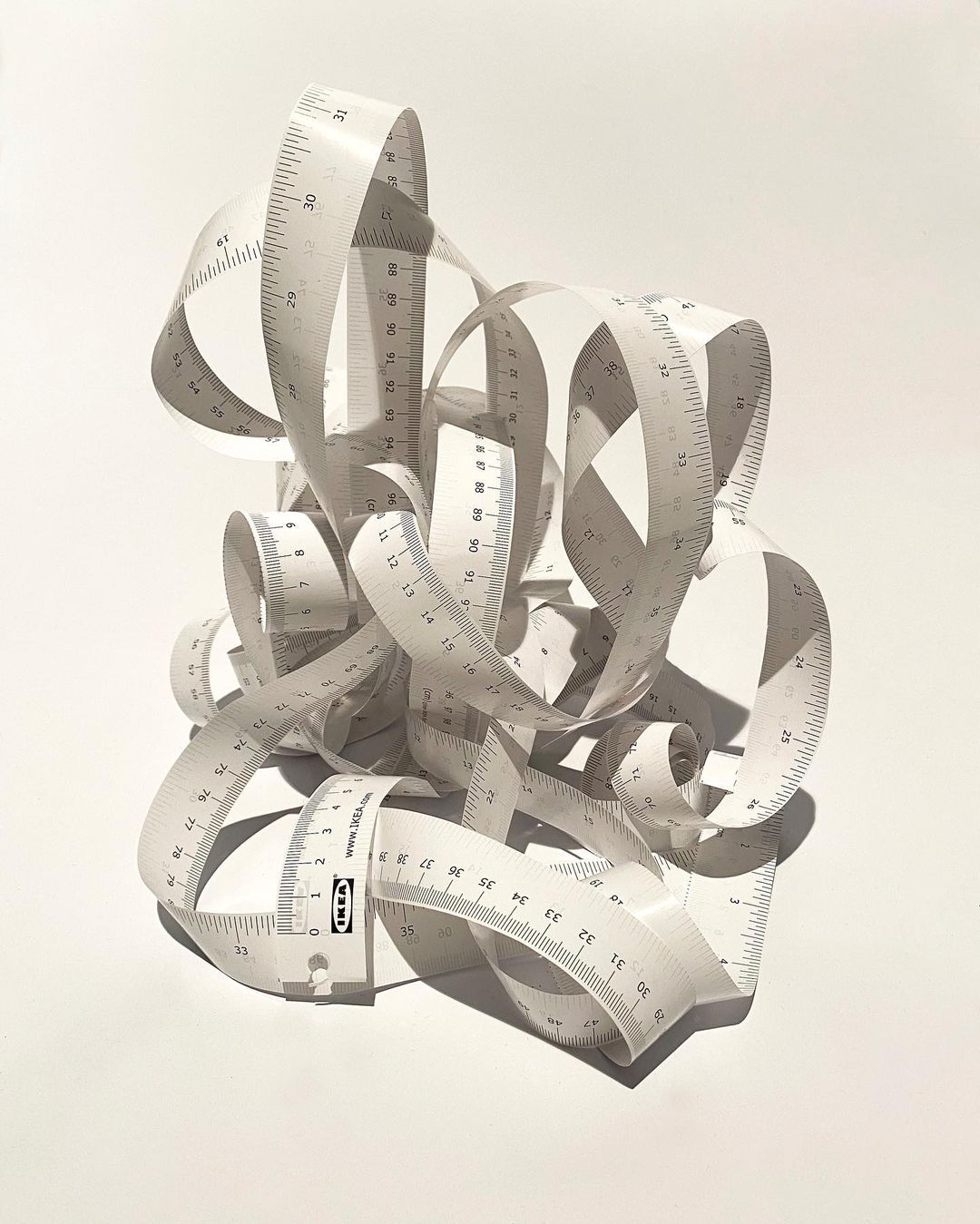 Close-up studio shot of a tangled mess of white measuring tapes with black markings, creating an abstract design on a white surface.