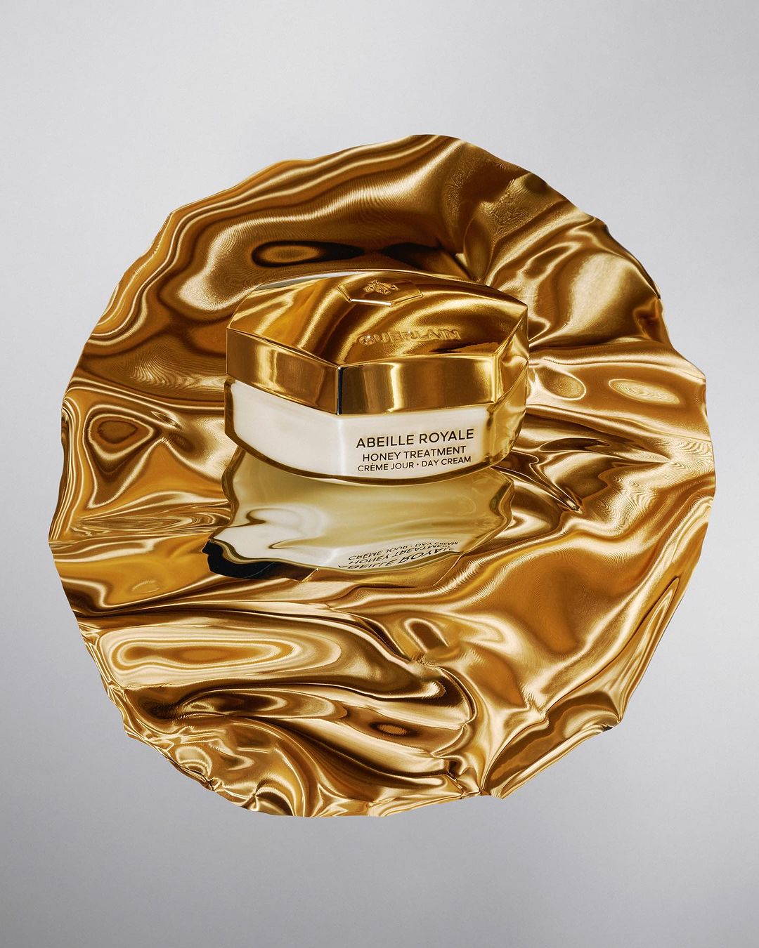 Close-up of Guerlain's Abeille Royale honey treatment cream jar with golden lid and white base, sitting atop a reflective gold fabric.