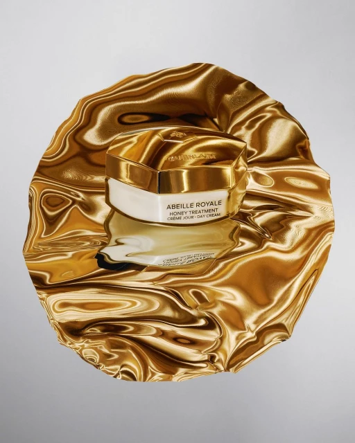 Close-up of Guerlain's Abeille Royale honey treatment cream jar with golden lid and white base, sitting atop a reflective gold fabric.