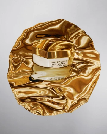 Close-up of Guerlain's Abeille Royale honey treatment cream jar with golden lid and white base, sitting atop a reflective gold fabric.