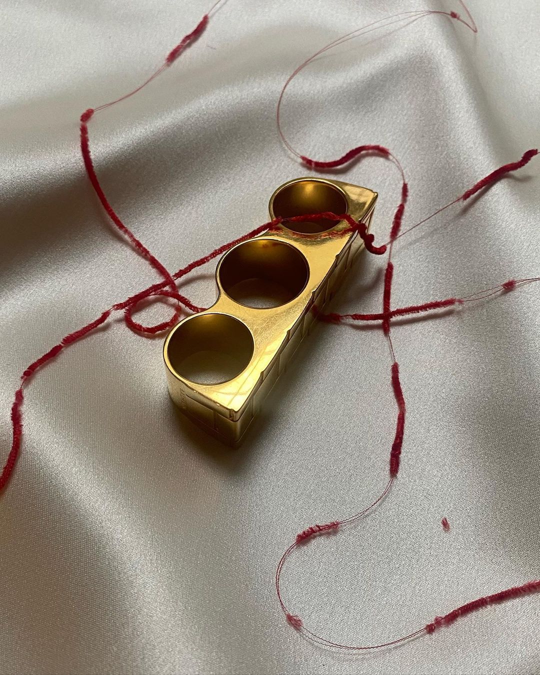 A gold three-finger ring is displayed on a silky white fabric, adorned with delicate, scattered red threads. Soft, diffused lighting.