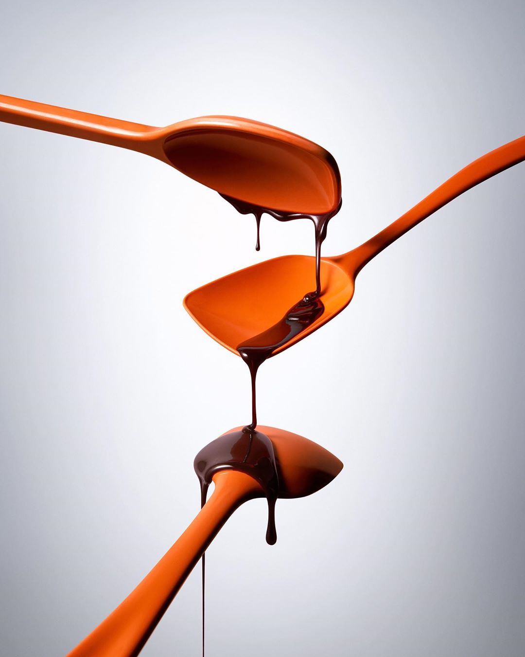 Three orange spoons are stacked vertically, with dark chocolate dripping from one spoon to the next against a light gray gradient background.