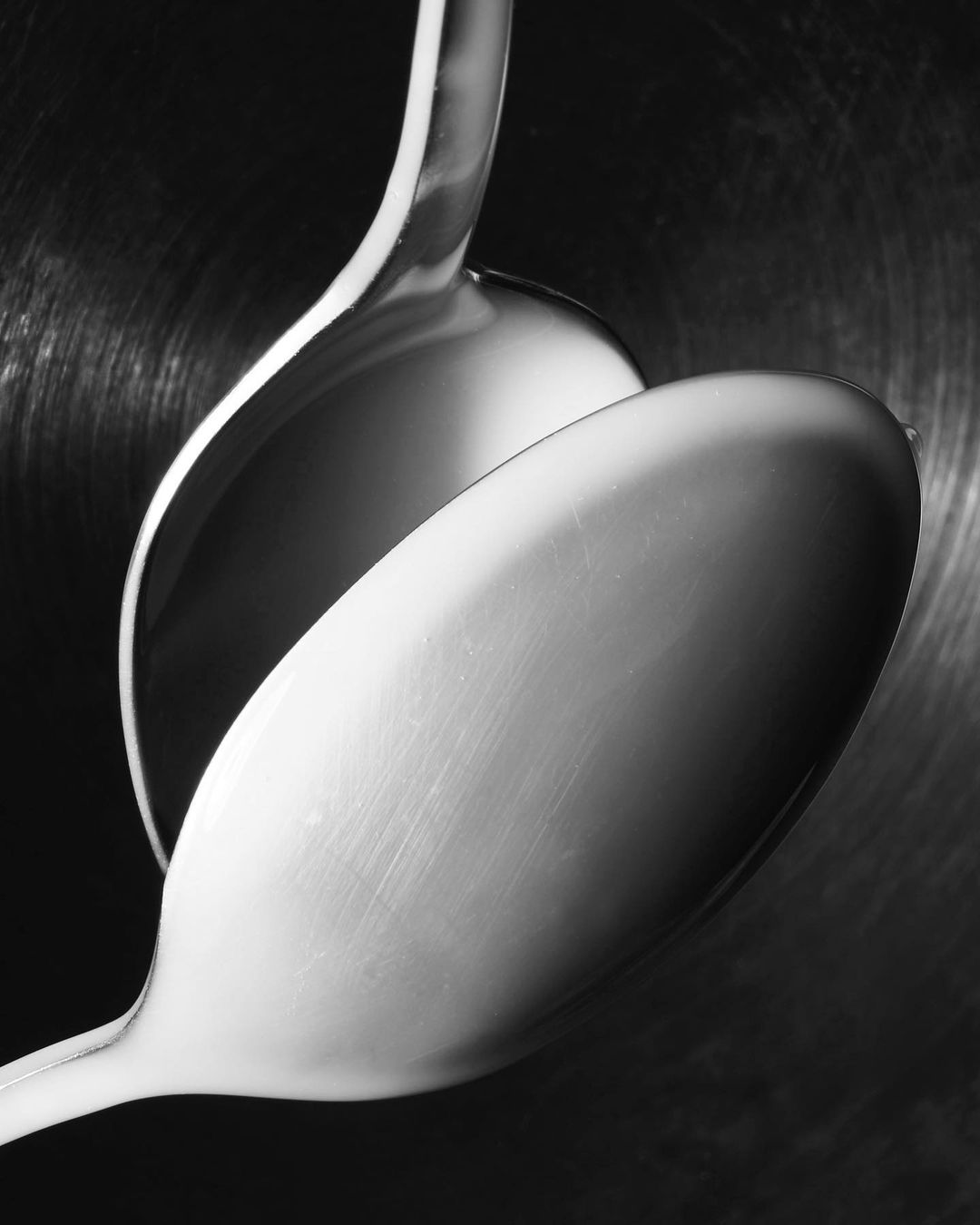 Monochromatic image of two spoons overlapping, creating an abstract composition of reflections, light, and shadows on metal.