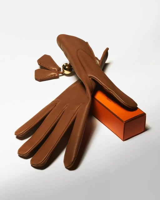 Pair of caramel-colored leather gloves arranged next to a small orange box, creating a minimalist and fashion-forward still life. 