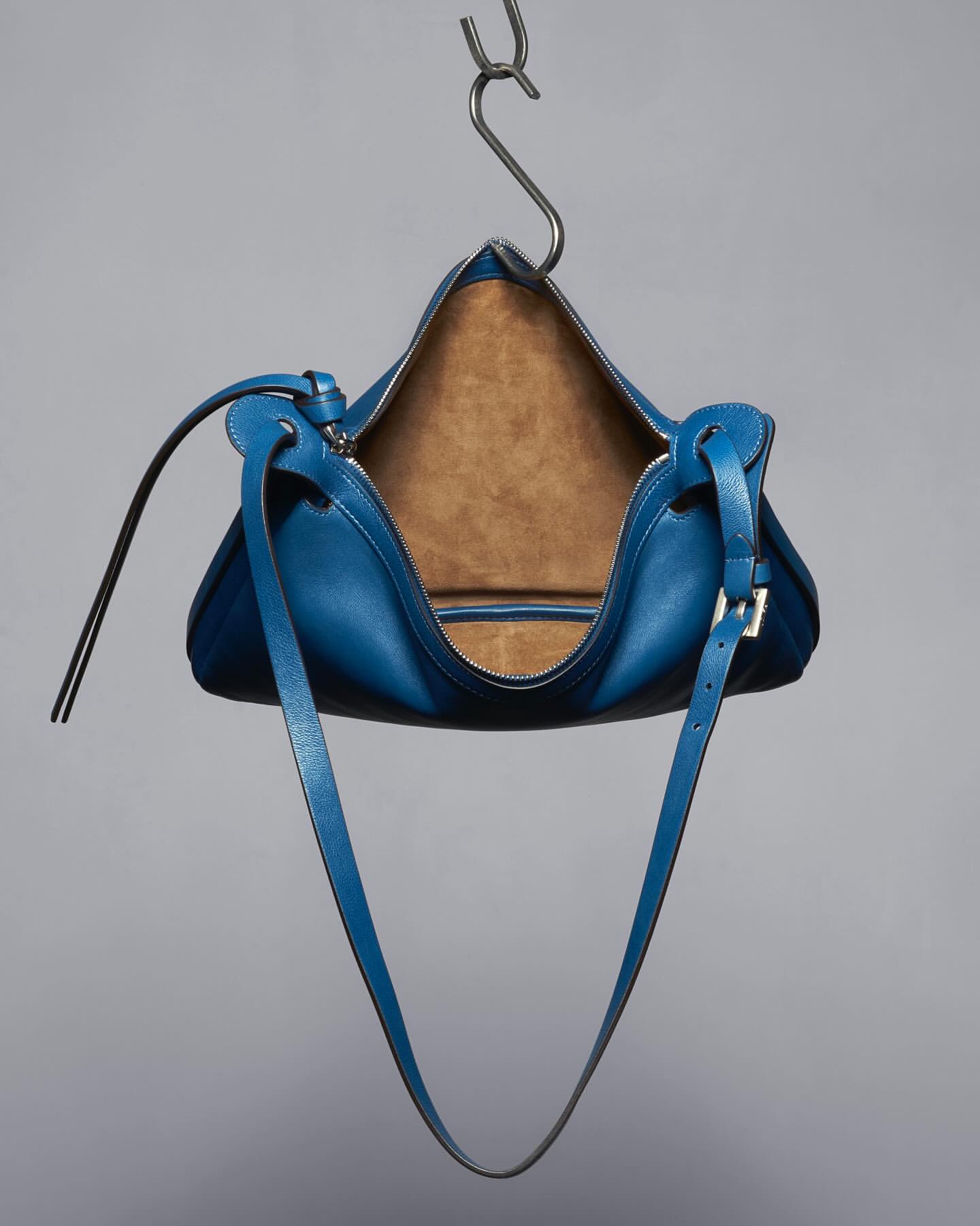 Studio shot of a modern blue leather handbag hanging from a silver hook against a gray background, showcasing its design and craftsmanship.