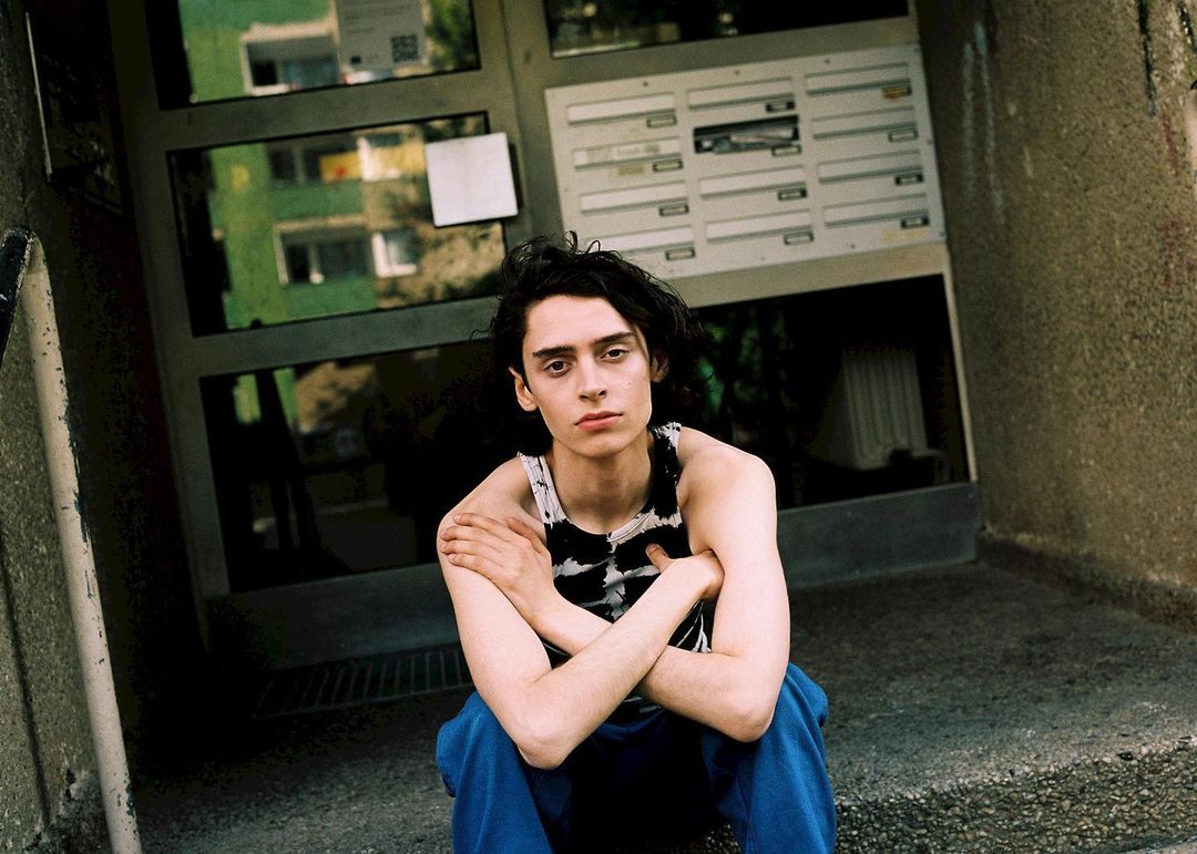 Young adult sits in an urban doorway wearing a black-and-white striped tank top and blue pants, looking pensively at the camera.