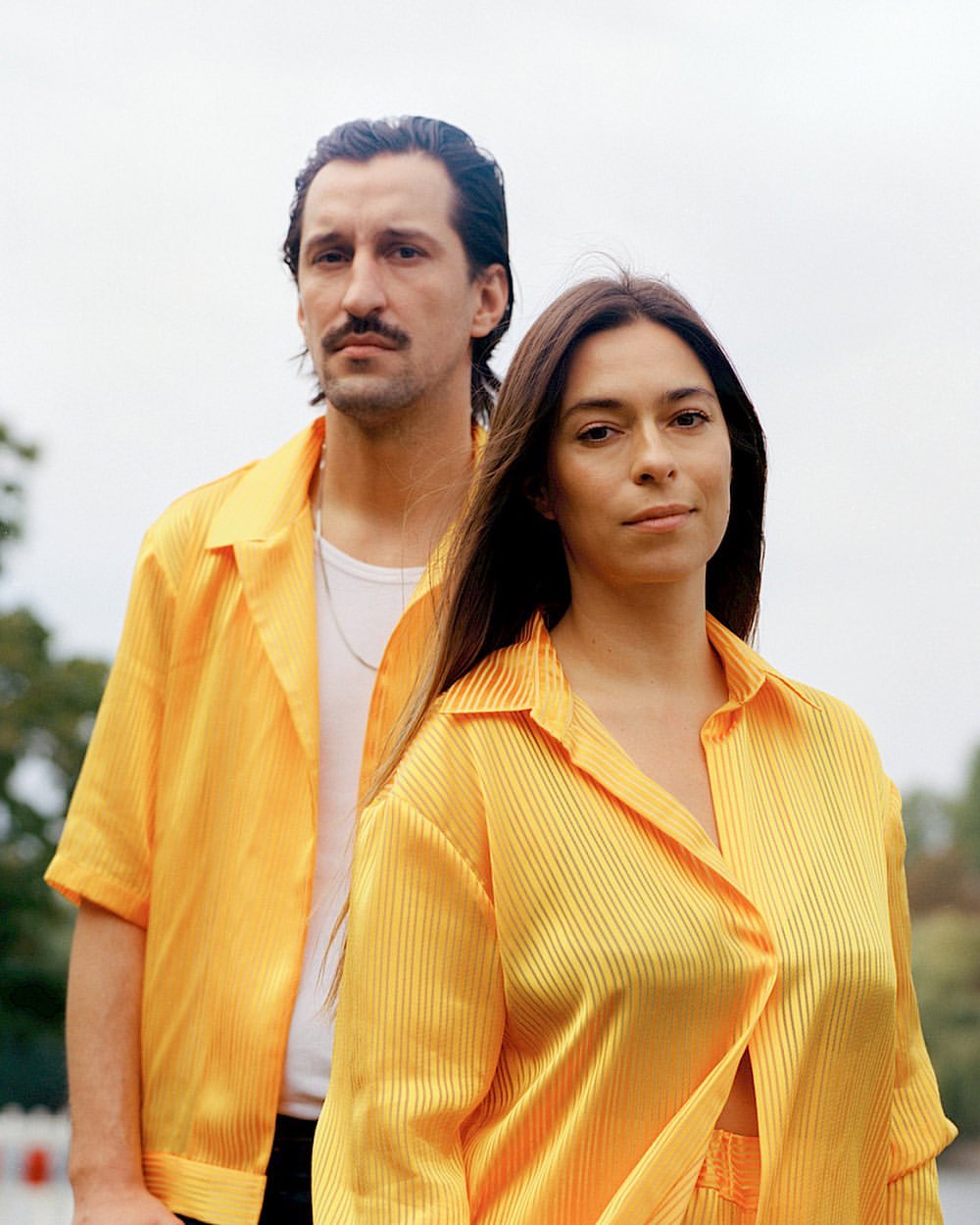 Stylish couple in matching yellow shirts pose outdoors against a blurred background. Fashionable, coordinated outfits, natural light.