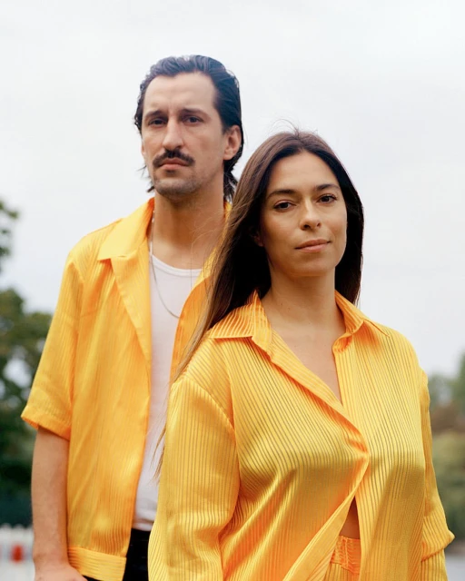 Stylish couple in matching yellow shirts pose outdoors against a blurred background. Fashionable, coordinated outfits, natural light.