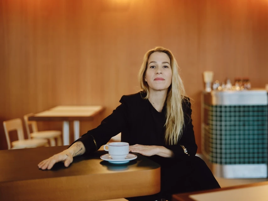 Adult woman sitting at a table with a coffee cup and saucer, in a restaurant with a warm, inviting atmosphere. 