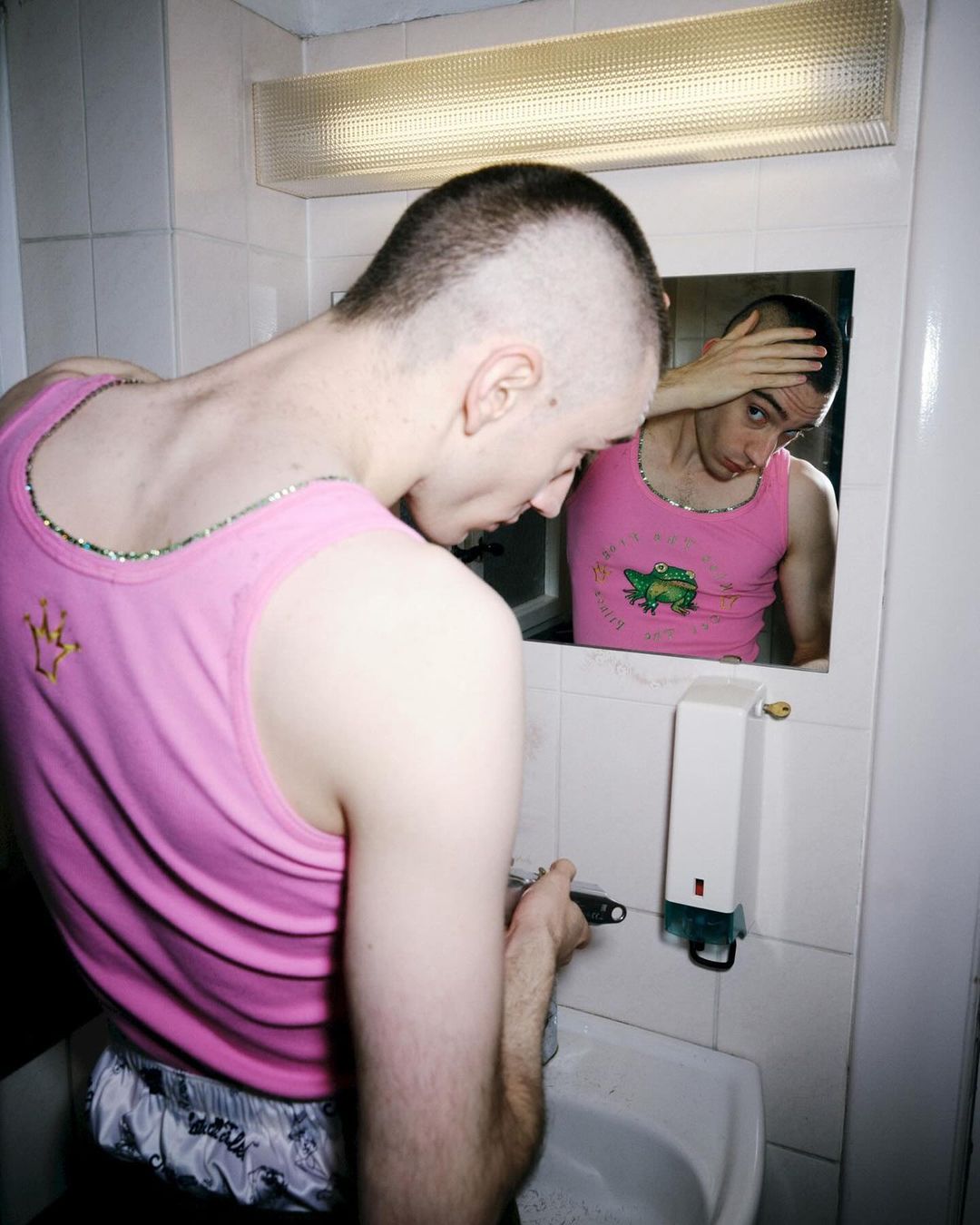 Man with a shaved head in a pink tank top shaves in a bathroom in front of a mirror in an indoor setting with cool lighting.