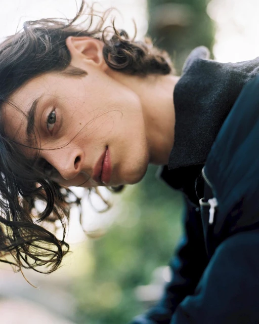 Portrait of a young adult with long brown wavy hair wearing a dark blue jacket, gazing into the camera in an outdoor setting.