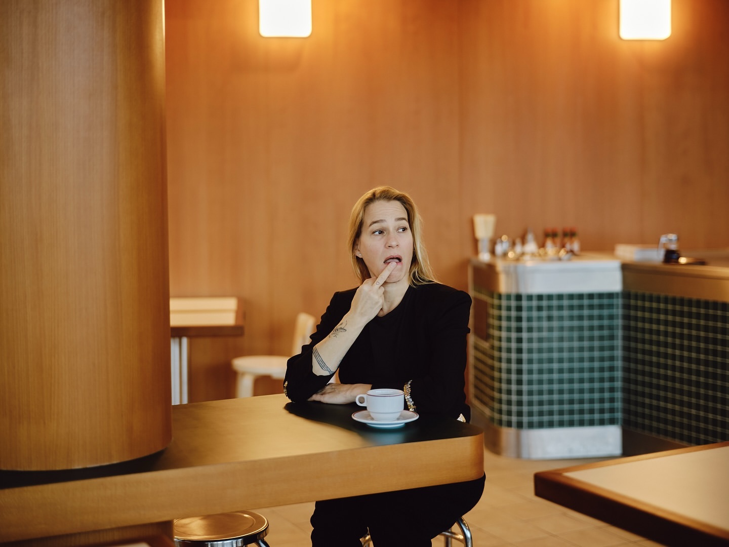 Woman sitting at a table in a cafe with a finger in her mouth. She is wearing a black jacket and there is a cup of coffee.