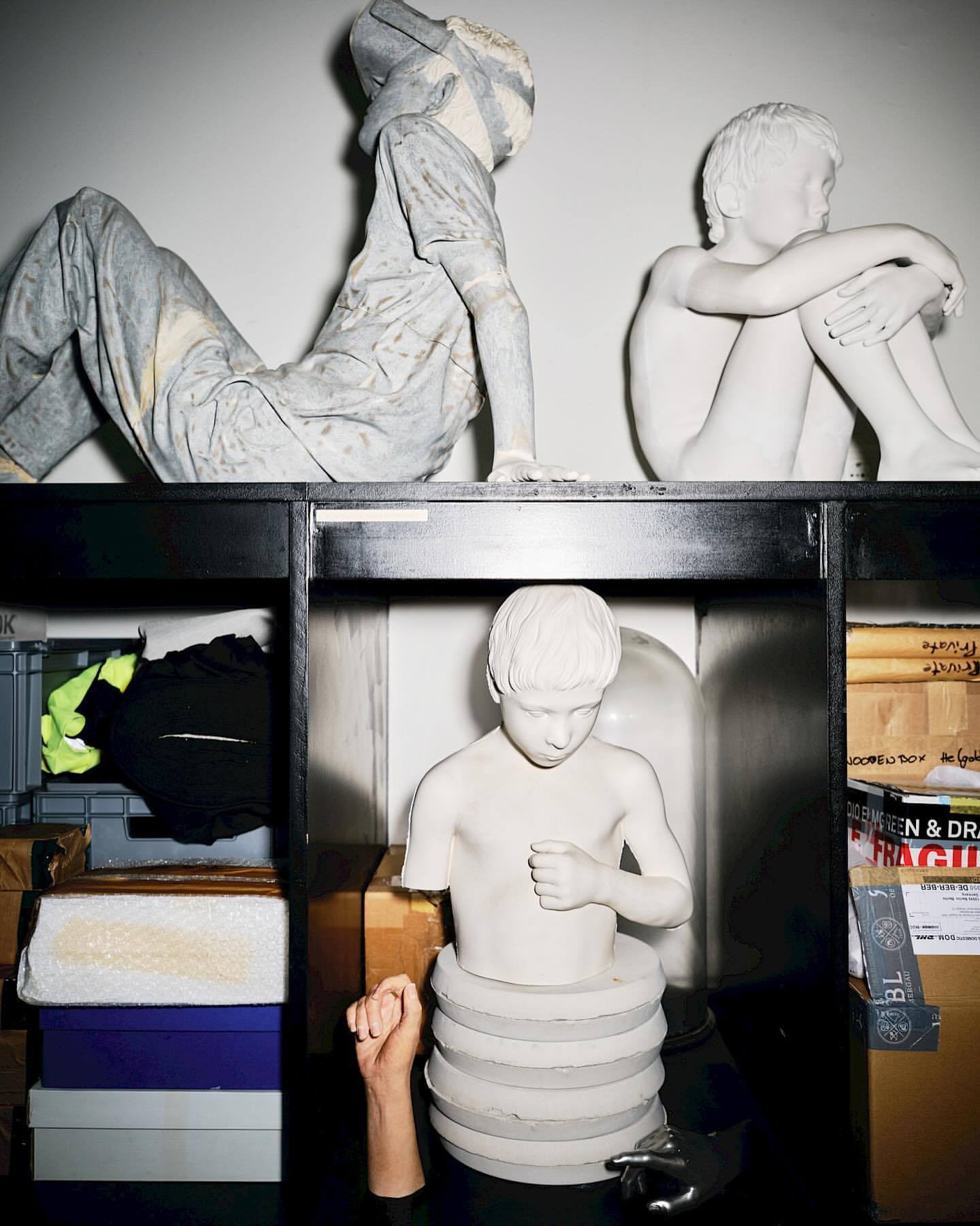 Art installation featuring a collection of white plaster sculpture busts and figures, set against a white background inside a black shelving unit.