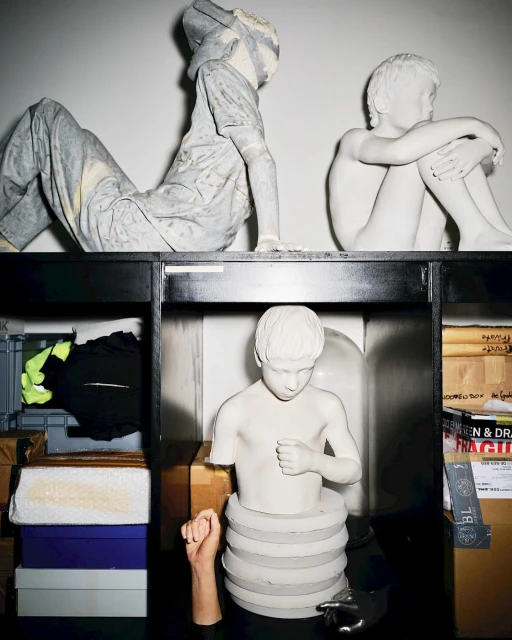 Art installation featuring a collection of white plaster sculpture busts and figures, set against a white background inside a black shelving unit.