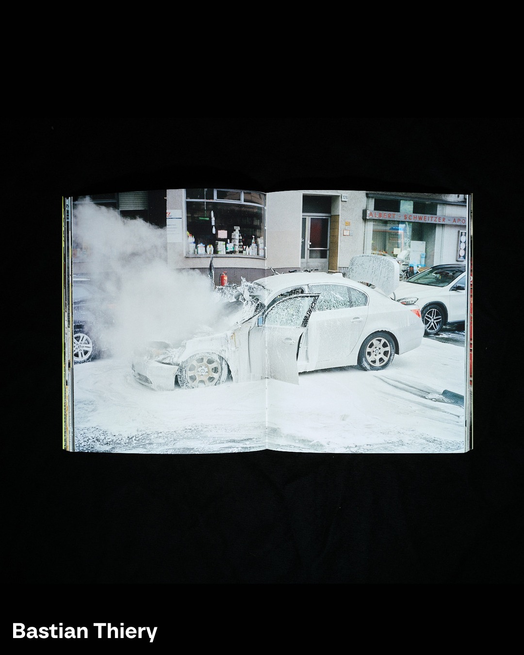 Open book page featuring a white car covered in what appears to be white foam in a city street setting, creating a surreal visual.