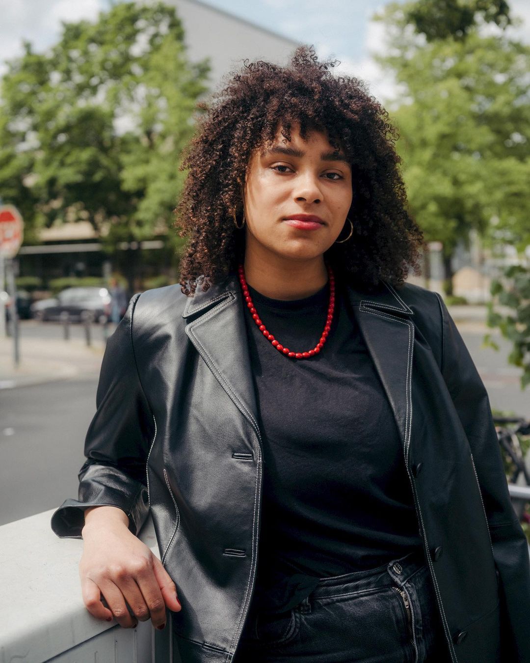 A woman with curly hair poses outdoors wearing a black jacket and red necklace. Trees and a building are visible in the background.