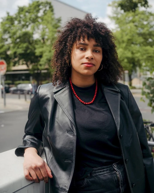 A woman with curly hair poses outdoors wearing a black jacket and red necklace. Trees and a building are visible in the background.