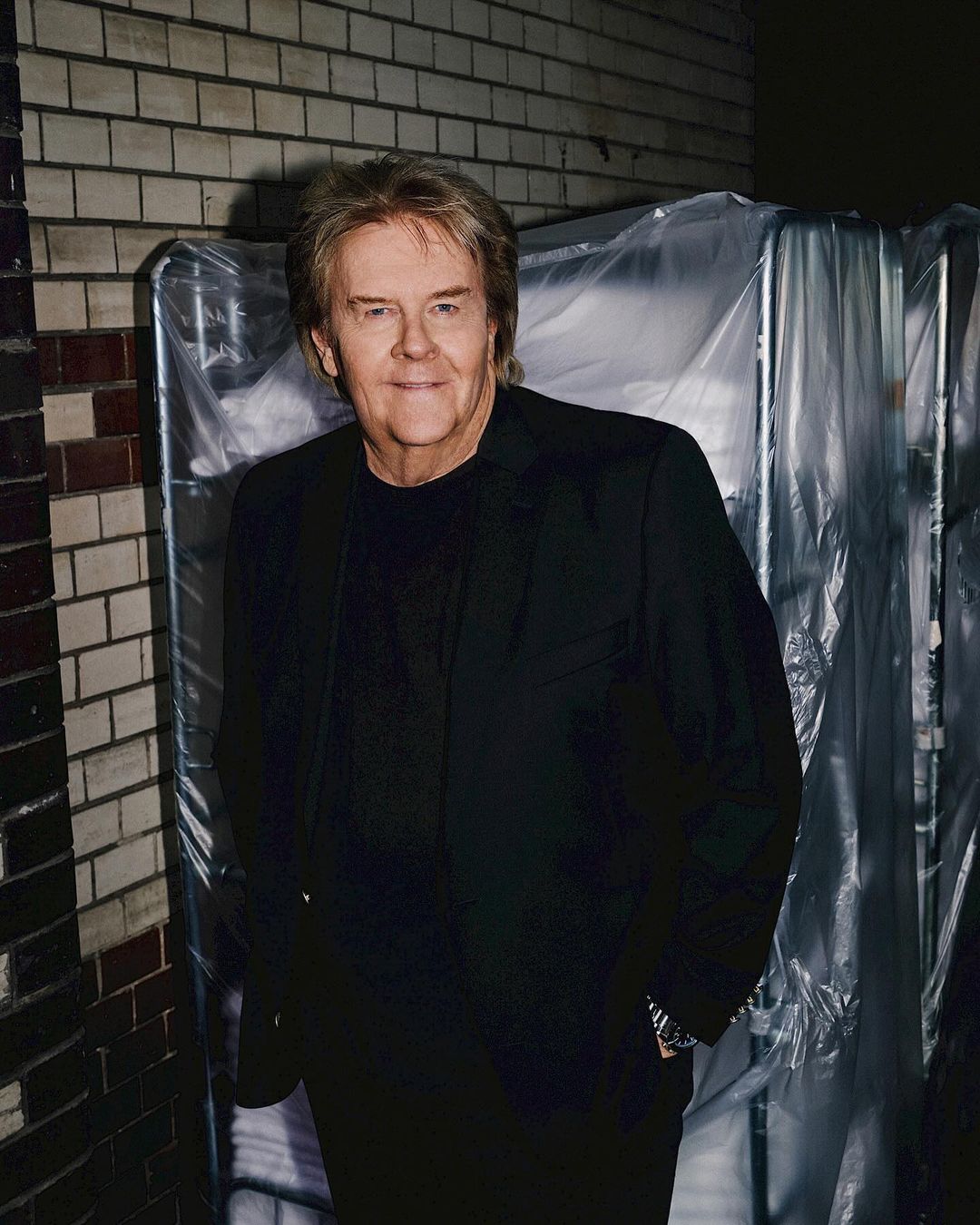 Portrait of an adult male with blond hair wearing a black blazer, posing against a brick wall and equipment covered in plastic sheeting.