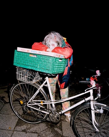 Person in costume leans on pizza boxes atop bicycle on a dark street, illuminated by artificial light, creating an eerie, surreal image.