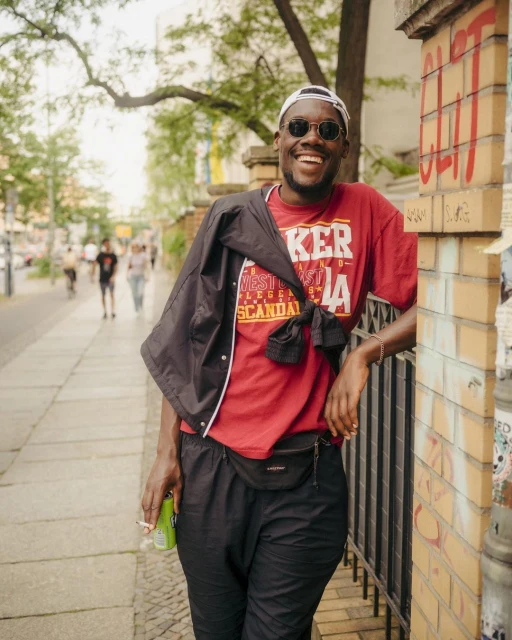 Smiling man stands near a brick wall outdoors. He wears sunglasses, a red t-shirt with text, dark pants, and a black jacket.