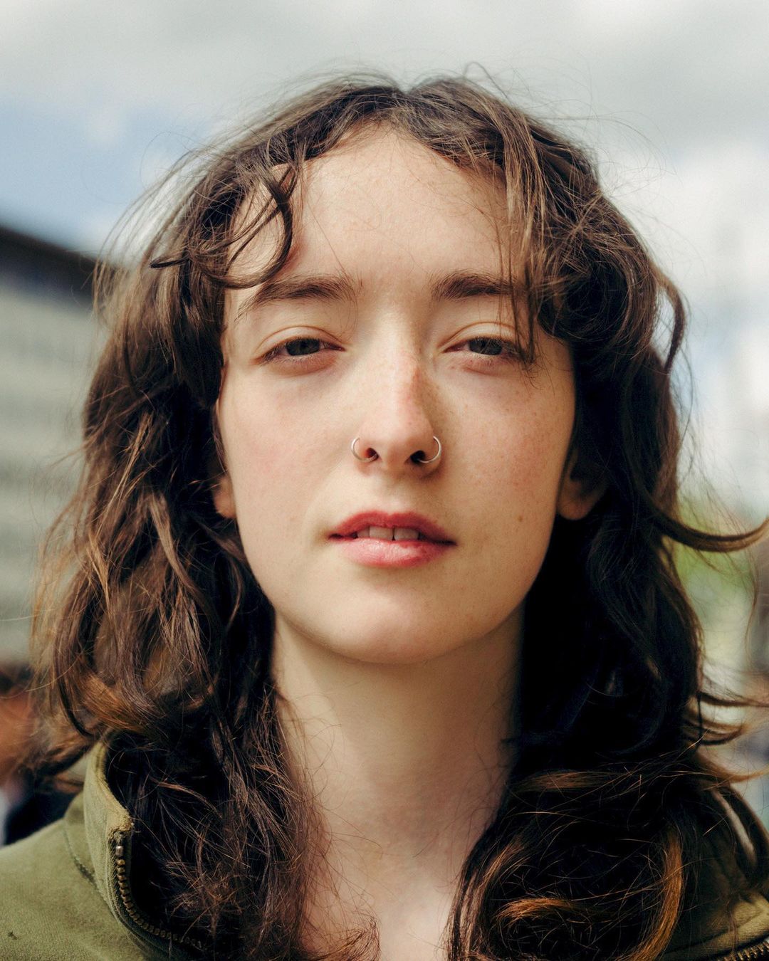 Close-up portrait of a young woman with wavy brown hair, septum ring, and olive green jacket against an out-of-focus background.